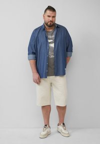 Blauwe denim overhemd met opgerolde mouwen, grijze graphic t-shirt, crèmekleurige shorts en witte sneakers met bruine accenten.
