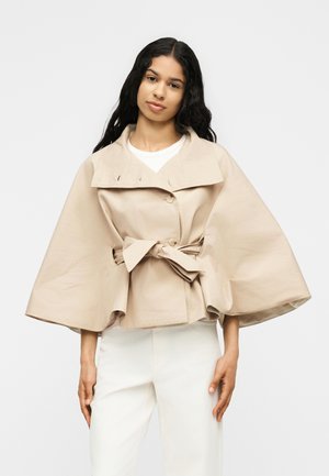 Femme aux longs cheveux foncés porte une veste beige style cape avec de larges manches et une ceinture, associée à un pantalon blanc, debout devant un fond uni.