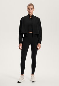 Černá zkrácená bunda s vysokým límcem, doplněná černým crop topem a legínami. Model má na sobě bílé sportovní boty. Hladký materiál, přiléhavý střih.