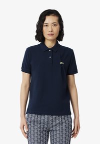 Tummansininen polo-paita, jossa on klassinen kaulus ja kolme nappia. Vasemmassa rinnassa pieni vihreä brodeerattu logo. Lyhyet hihat.