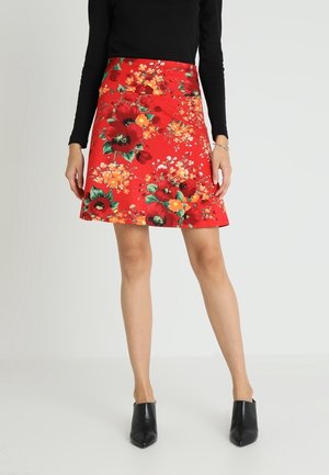 Mujer con falda roja con flores grandes, top negro de manga larga y botines negros con punta en pico, de pie sobre un fondo claro.