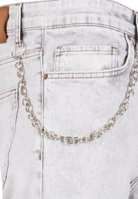 Hõbedane kett, mis on kinnitatud heledate hallide denim teksade külge, millel on nahast silt ja õmblemise detailid. Kett näib tekstuurne, koos omavahel seotud linkidega.