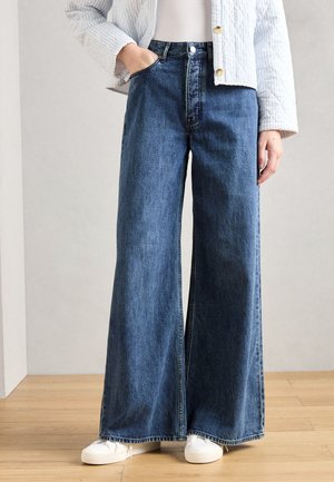 Jeans azuis de perna larga em denim com um design de cintura alta. Apresenta fecho com botão, bolsos frontais e uma bainha limpa. Combinados com ténis brancos.