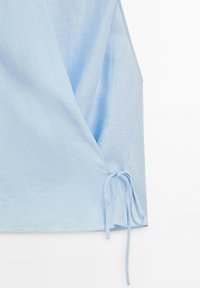 Haut cache-cœur sans manches bleu clair avec un lien en tissu sur le côté droit, fabriqué en tissu mélange de lin doux et respirant.