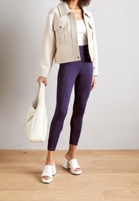 Beige zkrácená bunda se dvěma předními kapsami, bílý žebrovaný top, tmavě modré legíny a bílé muly na blokovém podpatku. Světlé pozadí a dřevěná podlaha.