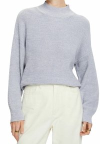 Lichtgrijze trui met een hoge hals, los gebreide structuur en verzakte schouders, gecombineerd met witte broek. Subtiele details op de manchetten.