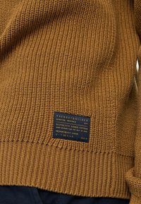 Hnědý žebrovaný pletený svetr s modrou textilní etiketou. Design se vyznačuje střední/ těžkou texturou a je inspirován námořními tématy.