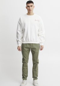 Sudadera blanca de cuello redondo con una ligera textura y detalles impresos en el pecho, combinada con pantalones verdes oliva y zapatillas blancas.