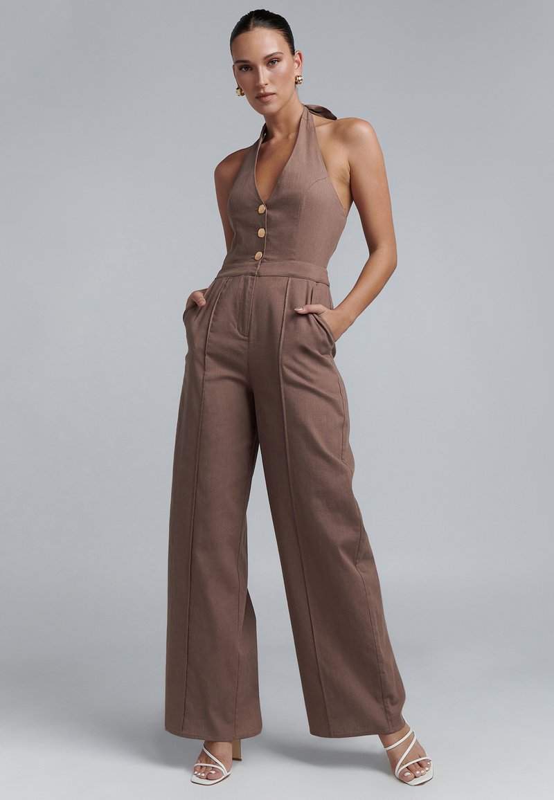 Bruine linnen jumpsuit met een halternek, knopen aan de voorkant, wijde pijpen en zijzakken. Glad textuur en minimalistisch design.