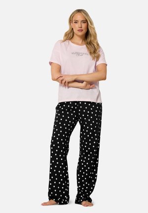Frau steht barfuß und trägt ein hellrosa T-Shirt mit dem Text "Sleepover Club" und schwarze Pyjamahosen mit Punkten vor weißem Hintergrund.