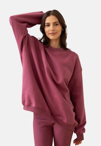 Kobieta z długimi ciemnymi włosami, ubrana w przesadnie dużą fioletową sweatshirt i pasujące spodnie, pozująca z uniesioną jedną ręką za głową.