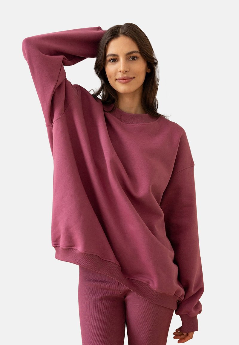 Kobieta z długimi ciemnymi włosami, ubrana w przesadnie dużą fioletową sweatshirt i pasujące spodnie, pozująca z uniesioną jedną ręką za głową.