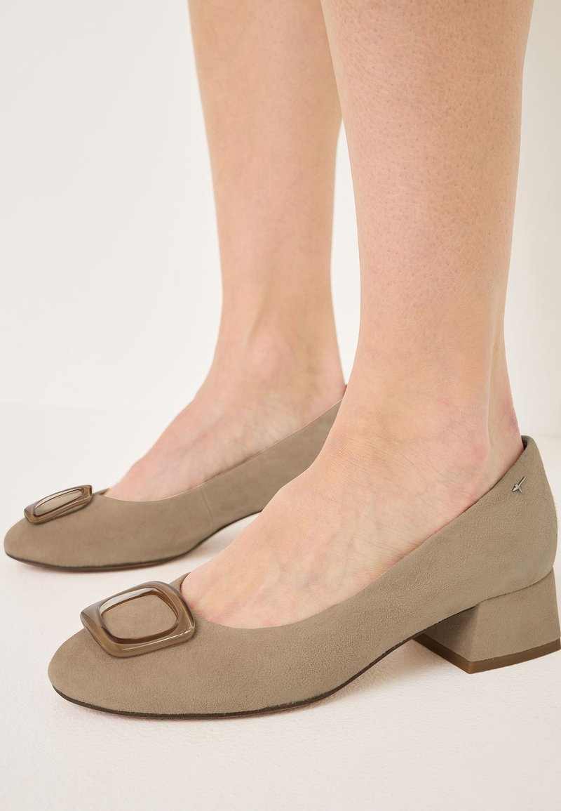 Pieds portant des chaussures à talon carré bas en daim beige avec une boucle carrée décorative sur le bout, sur un fond uni.