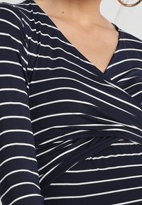 Mörkblå omlottblus med vita horisontella ränder. Har lång ärm och en tvistad frontdesign för en figurnära silhuett. Slät tygtextur.