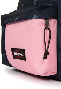 Mochila Eastpak con un tejido azul marino que presenta un pequeño patrón de árboles, un bolsillo frontal rosa, cordón ajustable negro y un parche con el logo.