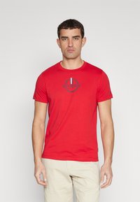 Rød kortærmet t-shirt lavet af bomuld. Har et marineblåt logo med et laurbærdesign og hvide striber ved halsen. Minimalistisk stil.