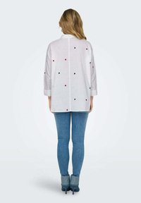 Camicia bianca a maniche lunghe con ricami a forma di cuore rossi, blu e rosa sparsi. Abbinata a jeans blu e tacchi grigi.