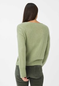Donna con lunghi capelli scuri che indossa un maglione lavorato a maglia verde chiaro e pantaloni verdi scuri, vista da dietro su uno sfondo semplice.