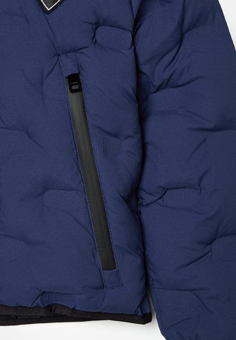 Dunkelblaue Daunenjacke mit gestepptem Futter, ausgestattet mit einer schwarzen Reißverschlusstasche und einem kontrastierenden Saum. Insgesamt weiche Textur.