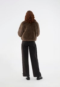 Chaqueta de pelo sintético marrón con un corte holgado, llevada sobre unos pantalones anchos de tiro alto en negro con detalles de botones en los laterales y zapatos negros.