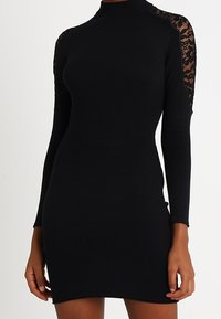 Schwarzes figurbetontes Kleid mit langen Ärmeln, hohem Ausschnitt und Spitze an den Schultern. Hergestellt aus einem glatten, elastischen Stoff.