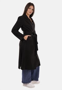 Donna che indossa un cappotto nero con cintura, jeans larghi blu e sneakers bianche, in piedi con una mano in tasca su uno sfondo bianco.