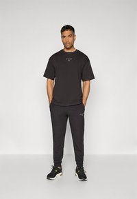 Melna īsroku T-krekls un atbilstoši sporta bikses, izgatavotas no mīksta auduma. Uz krūtīm ir nemanāma logotipa zīme, sporta biksēm kabatas, valkātas kopā ar melniem sporta apaviem.