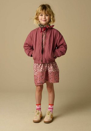 Kind lacht, draagt roze korte broek met patroon, een mauve gewatteerde jas, kleurrijke gestreepte sokken, beige sneakers en een sjaal met dierenprint en rode details.