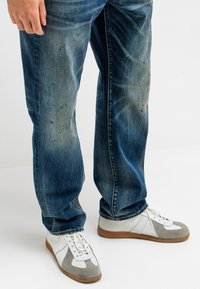 Blaue Jeans aus Denim mit einem ausgeblichenen Look und verwaschenen Details. Kombiniert mit weißen und grauen Sportschuhen mit hellbraunen Sohlen.