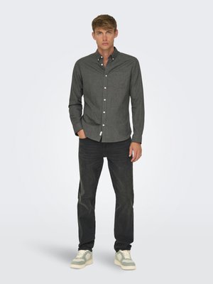 Jeune homme debout portant une chemise gris foncé boutonnée, un jean noir et des baskets gris clair sur un fond uni clair.