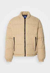 Beige puffatakki, jossa on korkea kaulus, vetoketjullinen etuosa, kaksi sivutaskua ja ribbireunat. Ominaisuuksina vaakasuora ompelukuosi ja logo rinnassa.