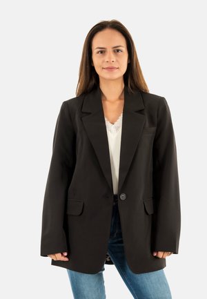 Femme aux longs cheveux bruns portant un blazer noir sur un top blanc et un jean bleu, debout devant un fond clair uni.
