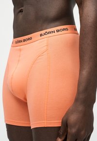 Κοντινό πλάνο ενός άνδρα με σκούρο δέρμα που φοράει μποξεράκια Björn Borg σε ροδακινί χρώμα, με λεπτομέρειες στη μέση και ραφές στον γοφό και τον μηρό του.