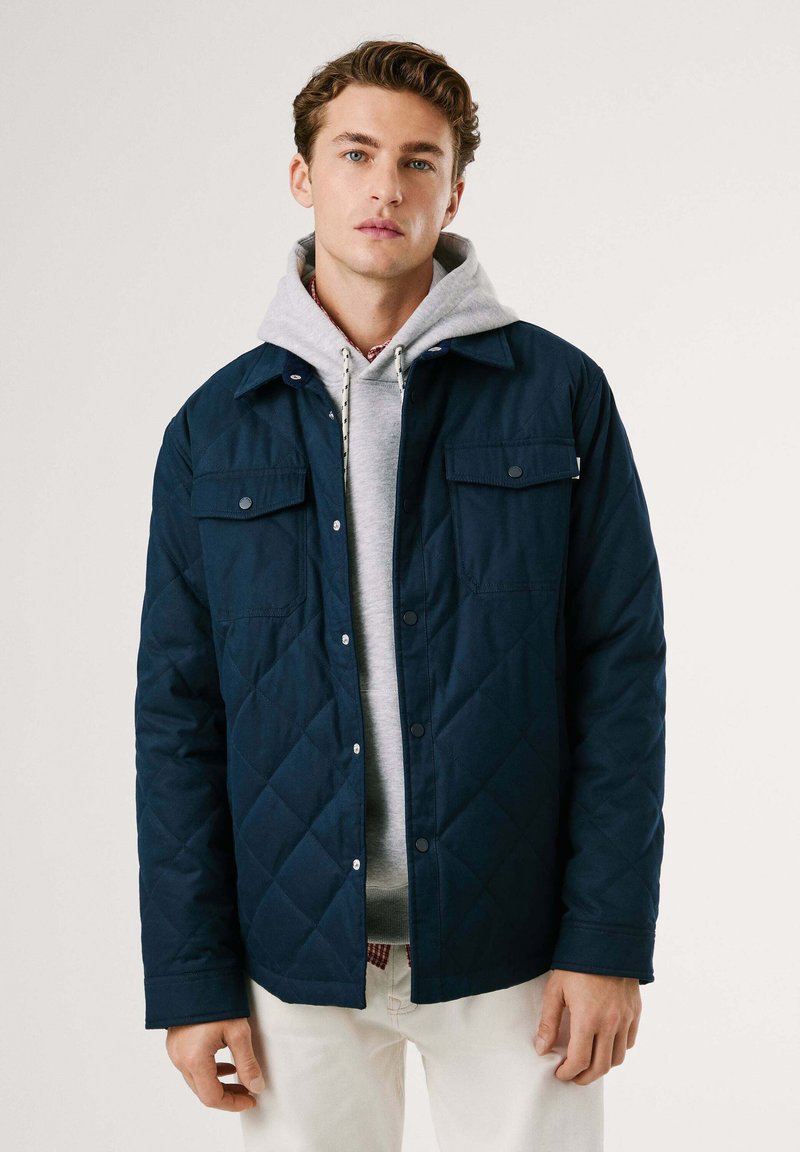 Chaqueta de punto azul marino con botones a presión, dos bolsillos en el pecho, superpuesta sobre una sudadera gris con capucha. Textura suave con diseño de patrón de diamantes.