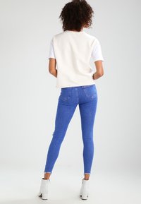 Top corto de manga corta blanco, que se lleva debajo de un chaleco crema, combinado con unos jeans ajustados de cintura alta azules y botas blancas hasta los tobillos.