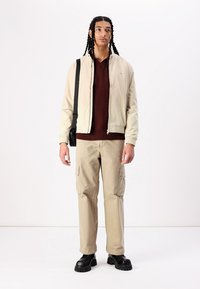 Blouson beige sur un pull côtelé bordeaux, associé à un pantalon cargo clair et des bottes noires épaisses ; le modèle pose avec un sac à épaule.