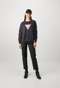 Sweat-shirt noir avec un logo triangulaire rouge portant "GUESS" et un point d'interrogation, assorti à un jean noir taille haute et des bottines noires.