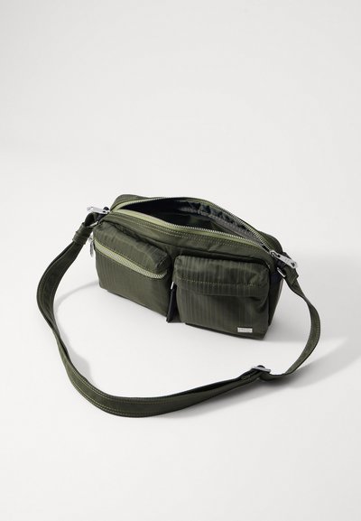 Grønn nylonveske med to frontlommepokker med glidelås, justerbar stropp og sølvhardware. Har et teksturert, stripet design.