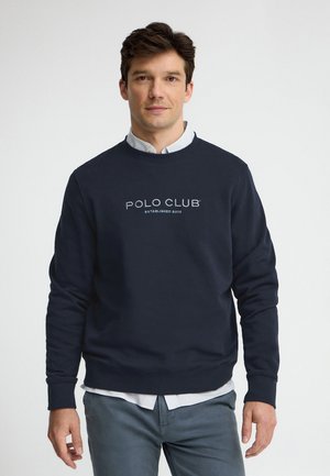 Uomo che indossa una felpa blu navy con il logo "POLO CLUB", camicia bianca con colletto sotto e pantaloni grigi, in piedi davanti a uno sfondo chiaro e uniforme.