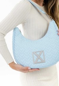 Femme portant une robe crème côtelée tient un sac à main matelassé bleu clair avec un logo géométrique argenté et une large bandoulière.