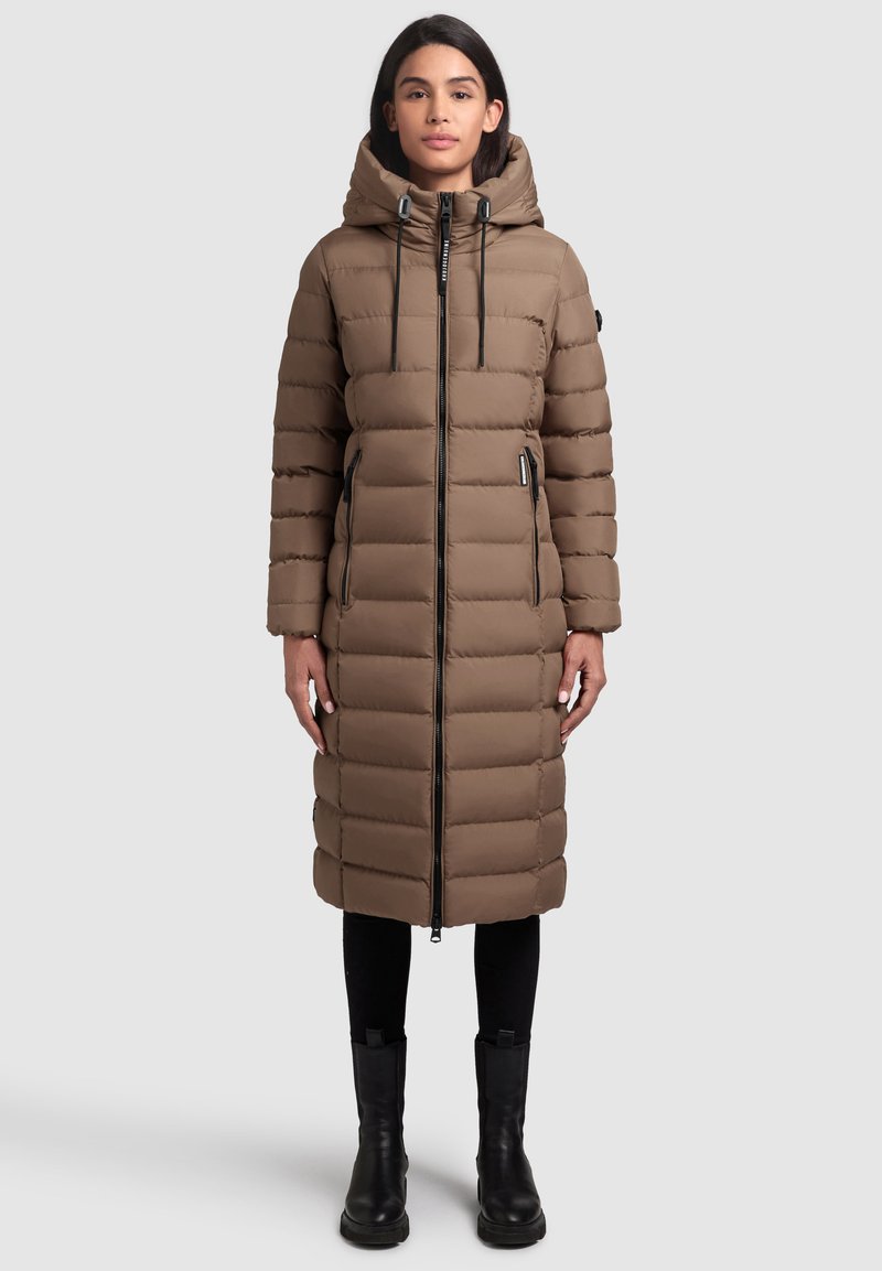 Bruine pufferjas met een capuchon, voorzien van een verticale gestikte patroon, een ritssluiting aan de voorkant en zwarte accenten. Gestyled met zwarte broek en enkelboots.