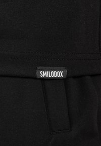 Czarna tkanina o gładkiej fakturze i płaskich szwach. Na krawędzi kieszeni widoczna jest mała biała metka z napisem "SMILODOX".