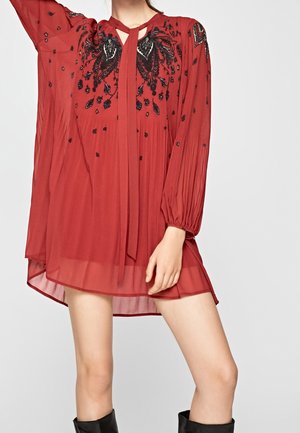 Czerwona sukienka z długim rękawem z czarną kwiatową haftem przy dekolcie, z półprzezroczystej tkaniny o plisowanej fakturze, z ozdobną kokardą przy szyi.