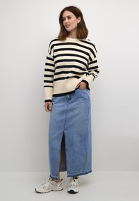 Pull rayé noir et blanc avec des poignets côtelés, associé à une jupe en denim bleue de mi-longueur avec une fente sur le devant, complétée par des baskets blanches.