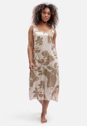 Donna con capelli ricci che indossa un vestito beige senza maniche con motivi floreali marroni ed elefanti, in piedi a piedi nudi su uno sfondo uniforme.