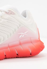 Chaussure de sport légère avec une tige blanche texturée, des accents roses et une semelle graduée du rouge vif au rose, ornée de rainures distinctives.