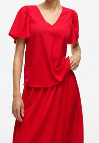 Czerwony top z dekoltem w serek, krótkimi bufkowatymi rękawami i luźnym krojem, wykonany z lekkiego materiału. Dół ma marszczony, zwiewny fason.