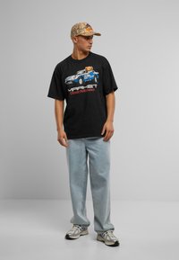 Svart oversized t-skjorte med grafisk trykk som viser en bil og en bjørn, kombinert med lyseblå jeans og et mønstrete cap. Sneakers med sølvdetaljer.