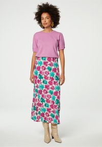 Lila kurzärmliges Oberteil, kombiniert mit einem langen, floralen Rock, der rosa und grüne Muster aufweist. Das Model trägt beigefarbene Knöchelschuhe mit quadratischen Absätzen.