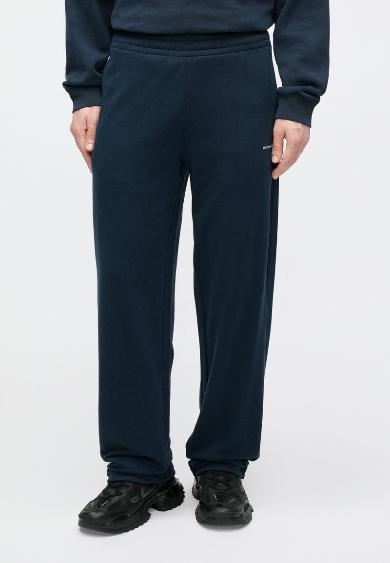 Pantalon de survêtement bleu marine en tissu doux, avec une taille élastique et des poches latérales, associé à des chaussures de sport noires.